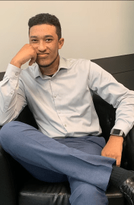 A young man dressed in a light-colored dress shirt and dark pants sits casually on a black leather chair, resting his chin on his hand with a thoughtful expression.